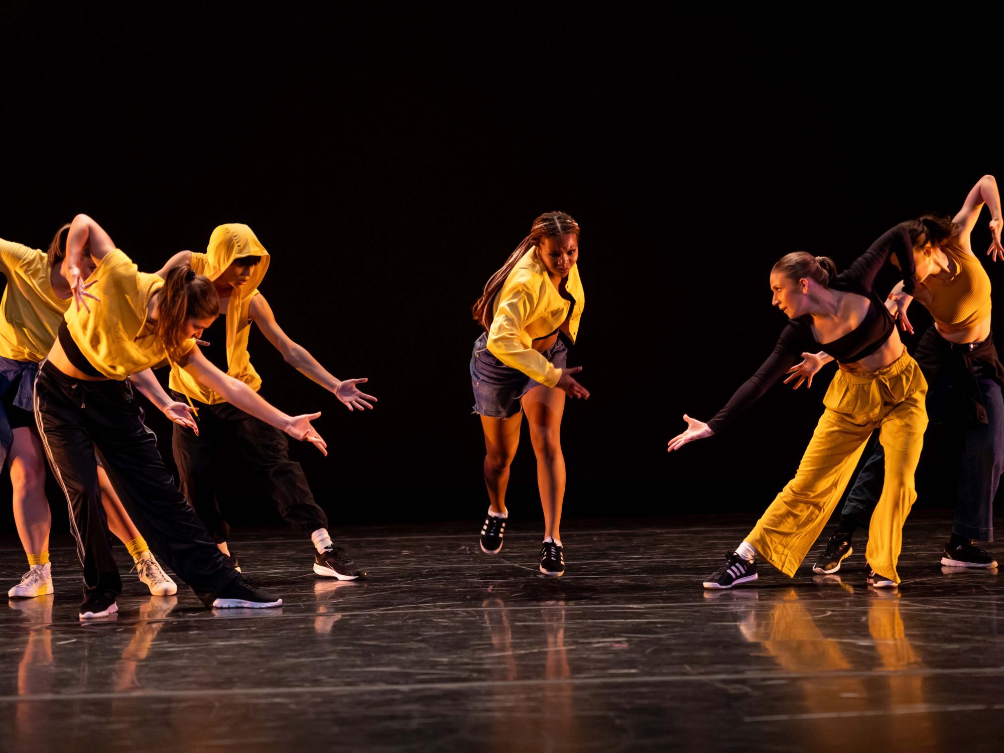 Dancers cloaked in black and yellow in Lone. Lean. Less. lean towards the center of the stage with their arms in a diagonal.