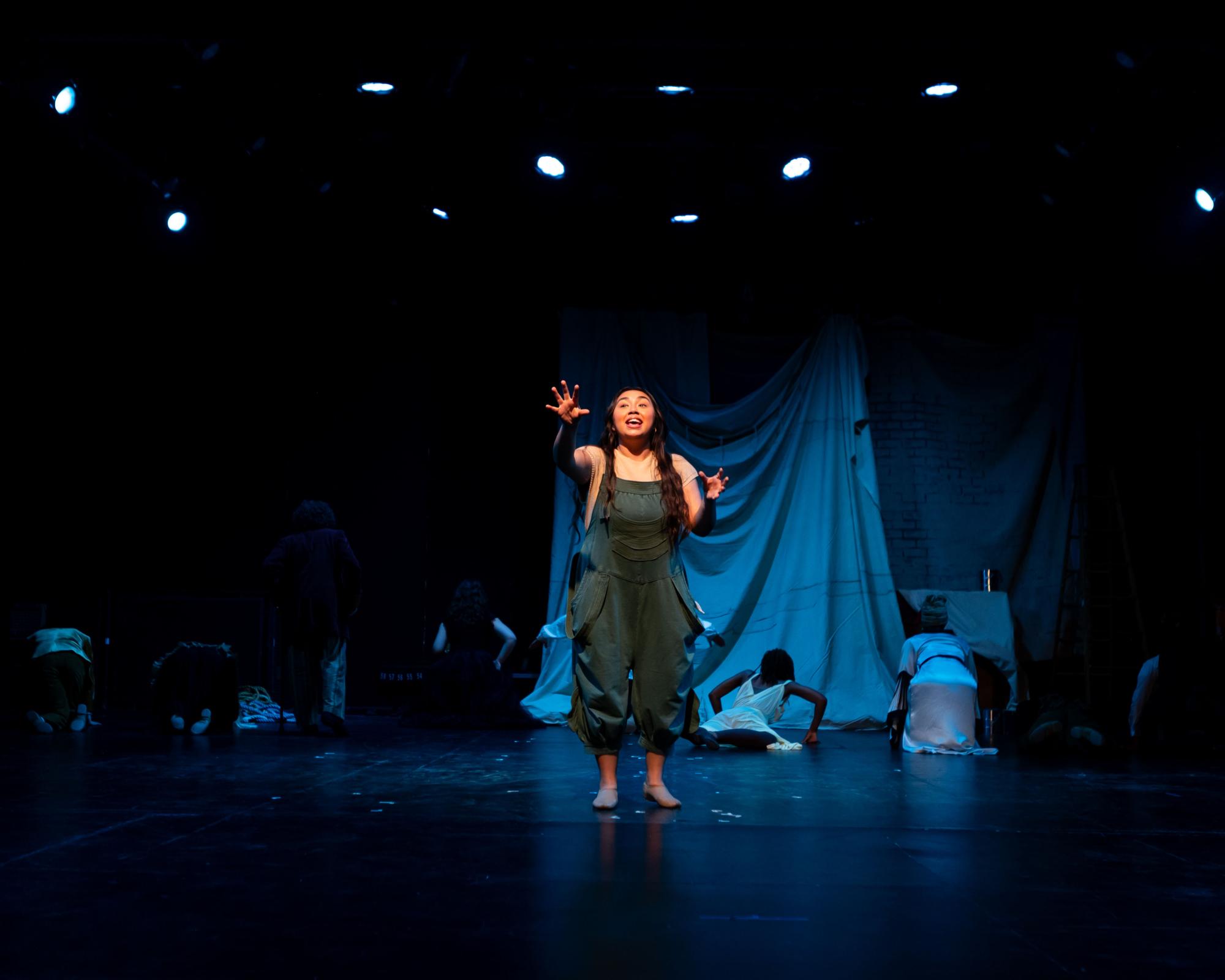 Yakov, played by Esmeralda Trevino, stands centerstage while the rest of the cast turns into moss growing up the back wall behind them. They reach out to the audience.