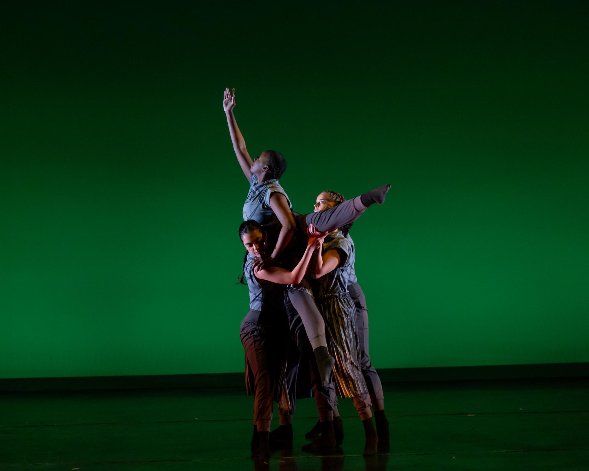 Photo of dancer being lifted by two other dancers in front of a green lit background