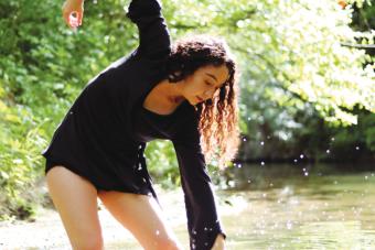 woman in all black with wavy brown hair dances in a shallow stream