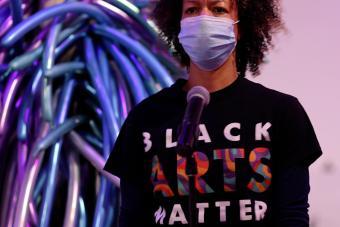woman with curly brown hair wearing a mask and a shirt that says "Black Arts Matters" standing in front of a microphone