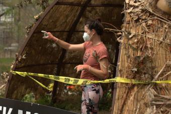 woman with mask and gardening gloves on points, standing within the Creek Monster Habitat art installation, a giant, nest-like structure shaped like a dome