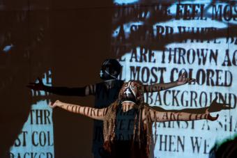 woman and man in all black with black masks on look up with outstretched arms, covered with projections of words and the silhouette of a tree