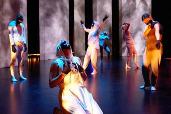 dance ensemble members wearing white hoodies and black masks pose, one on their knees, one with their arms in the air, two looking down.
