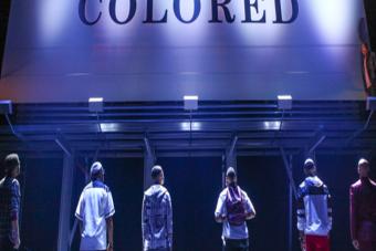 six Black men look up at a building that has the word "Colored" written in large letters on the front of it