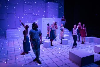 a group of actors walk around the Brockett Theatre, which is washed in blue, star-studded light