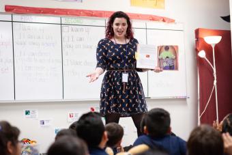 student teacher in a navy blue dress reads a book in front of a classroom of kids
