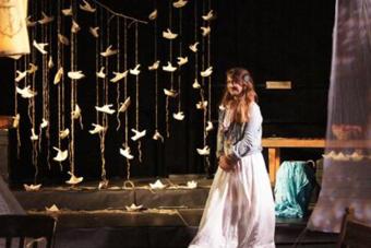 actress in a white dress stands in front of a collection of paper birds hanging from strings