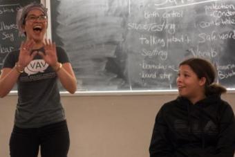 a woman stands in front of a blackboard with her hands out, while an onlooker watches