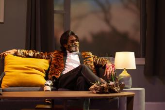 actor in a tiger striped suit coat and mask sits on a couch