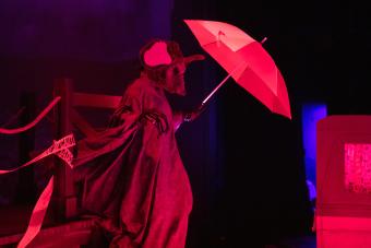 actor wearing a rat mascot head holds an umbrella, surrounded by red stage lighting