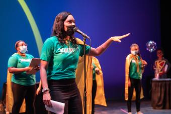 woman in a green shirt and yellow cape speaks into a microphone, with her left hand extended out