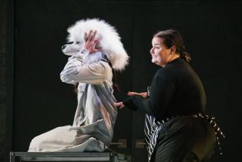 actor wearing a white dog head puts her hands on her ears, carted in by an actor in a striped apron