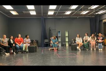 the cast for IN SISTERS WE TRUST rehearses a scene in which they're all sitting in a line of chairs