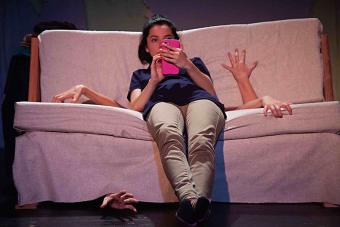 A scene from YAMEL CUCUY featuring an actress on her phone sitting on a couch with four hands reaching out from between the cushions