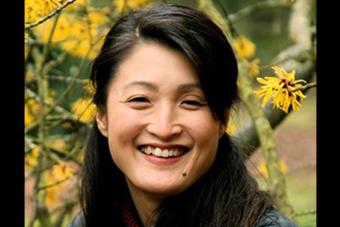 headshot for award-winning playwright Kimber Lee, smiling in front of yellow flowers