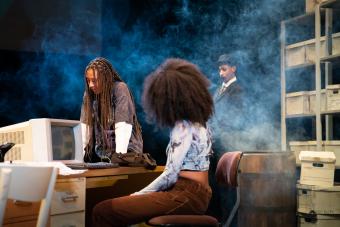 An actor looks down, leaning with their hands on top of a desk, while another actor looks on and a third actor watches from behind a wall of haze