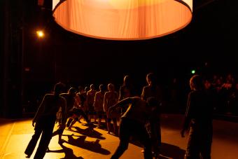 Cloaked in darkness, standing against a dimmed light, dancers are lined up as some move in and out of the diagonal they are standing in.
