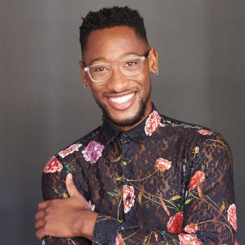 man with short black hair wearing a lacy floral shirt, glasses and silver hoop earrings