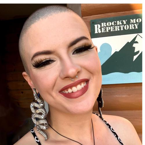 This person is wearing makeup and is sporting a shaved head. They are smiling at the camera, and they have a pale complexion.
