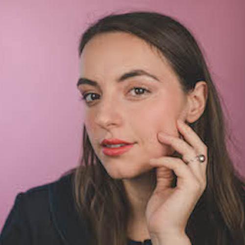 woman with brown hair places her left hand against her face, posing in front of a pink wall