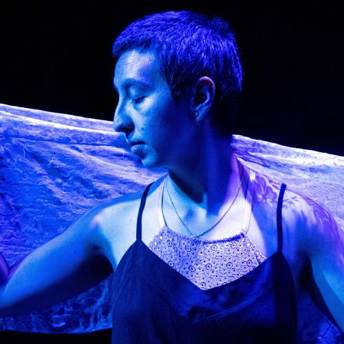 artist with short, dark hair faces their head to their right, holding a lacy cloth behind them and closing their eyes, all washed in a purple light